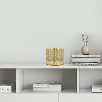 "A brass table lamp with a perforated metal body sitting on a white bookshelf next to a stack of books."