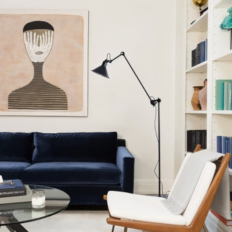 "Adjustable black floor lamp with angular arm and conical shade, placed beside a white armchair and a navy velvet sofa, with abstract art on the wall and bookshelves in the background."