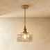 Brass pendant light with clear textured glass cylinder shade hanging against white textured wall with wooden headboard and dr