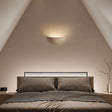 "Sleek, minimalist, half-moon shaped trimless light made of gypsum and metal, recessed into a bedroom wall, providing a soft, ambient glow above a modern bed with built-in headboard lighting."