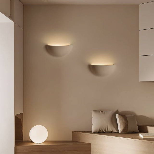 "Two sleek, minimalist, half-moon shaped trimless lights made of gypsum and metal, recessed into a living room wall, providing a soft, ambient glow above a seating area with a round table lamp."