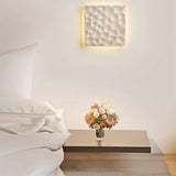 "A square travertine wall lamp with a textured surface, illuminating a bedroom wall above a bedside table with flowers."