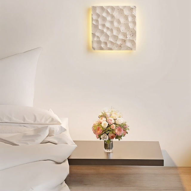 "A square travertine wall lamp with a textured surface, illuminating a bedroom wall above a bedside table with flowers."