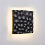 "Close-up of a black travertine wall lamp showing the hexagonal pattern and backlit glow on a textured wall."