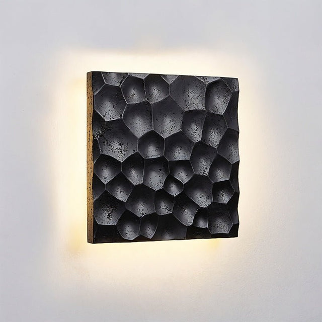 "Close-up of a black travertine wall lamp showing the hexagonal pattern and backlit glow on a textured wall."