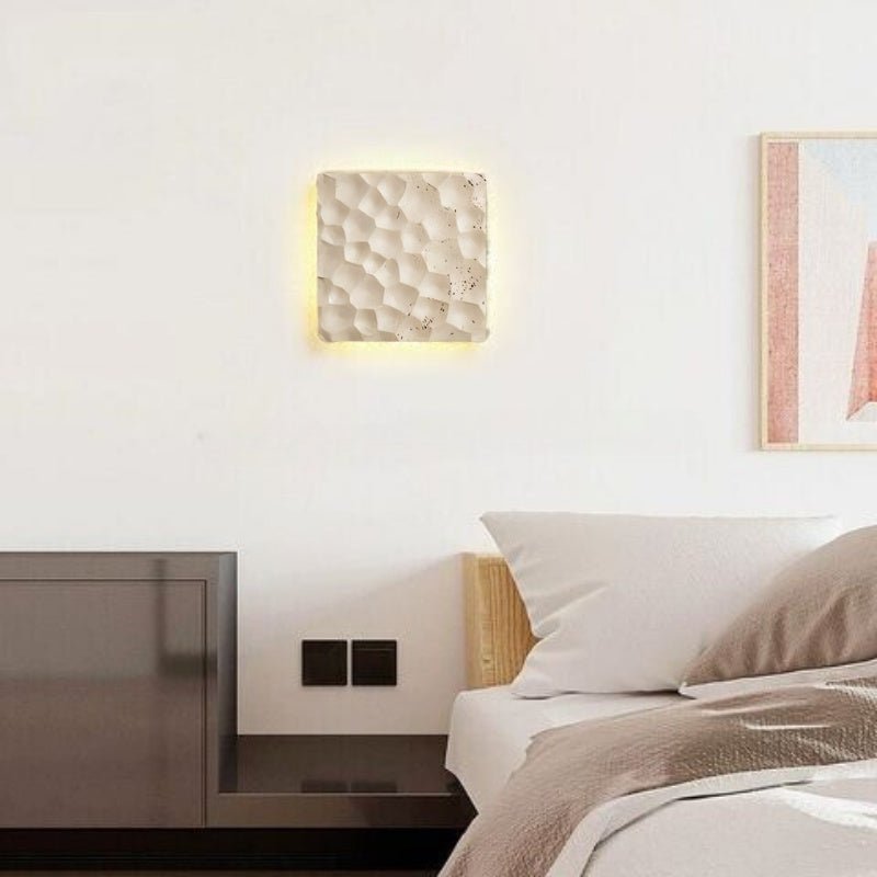 "A square travertine wall lamp with a textured surface, mounted on a white wall above a bedside table, emitting a soft glow."