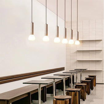 "Multiple cylindrical travertine pendant lights with frosted glass diffusers, hanging above tables and stools in a cafe."