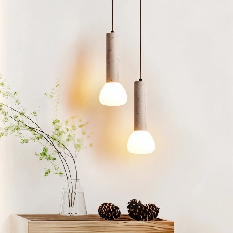 "Two cylindrical travertine pendant lights with frosted glass diffusers, hanging above a wooden cabinet with decorative items."