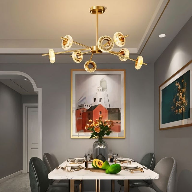 "A modern chandelier with circular glass accents and a gold metal frame hanging above a dining table with framed artwork."