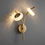 "A modern wall lamp with two lit textured glass shades and a gold metal frame mounted on a gray wall."