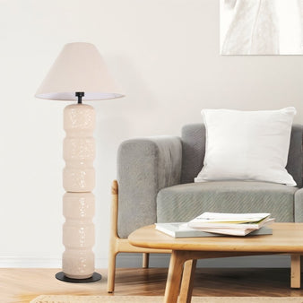 "A tall, beige ceramic floor lamp with a sculpted, segmented base and a conical fabric shade, standing next to a gray sofa with a white pillow and a round wooden coffee table in a modern living room."