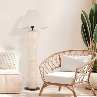 "A tall, beige ceramic floor lamp with a sculpted, segmented base and a conical fabric shade, standing next to a wicker armchair with a white pillow and a potted plant in a modern living room."