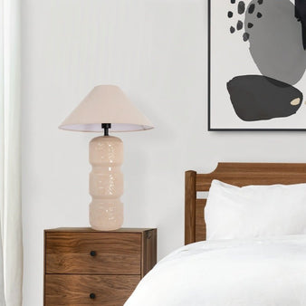 "A beige ceramic table lamp with a layered design, etched patterns, and a neutral fabric shade on a wooden nightstand in a modern bedroom with a white bed and a framed abstract art print."