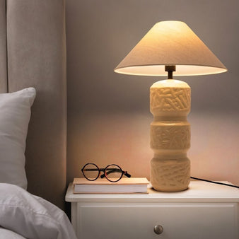 "A beige ceramic table lamp with a layered design, etched patterns, and a neutral fabric shade on a white nightstand in a bedroom, with a book and glasses next to it."