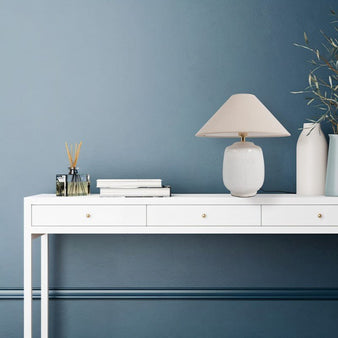 "A modern table lamp with a textured ceramic base and a tapered fabric lampshade on a white console table with books, a glass bottle with reeds, and a white vase with branches, against a blue wall."