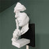 White sculptural face wall lamp with hand gesture mounted on black bracket against green wall.