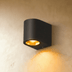 Dark gray cylindrical wall sconce with single light beam on white wall, lifestyle setting.