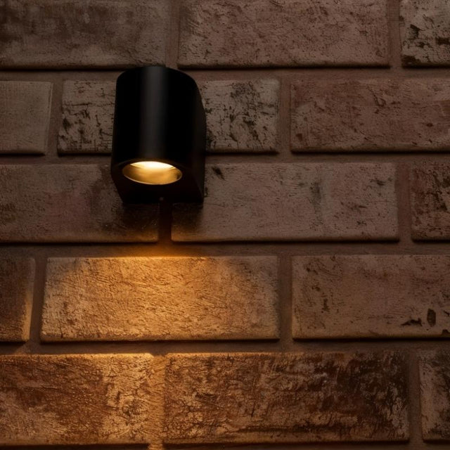 "Modern outdoor wall lamp with a dark aluminum body and glass diffuser, mounted on a brick wall, casting warm light."