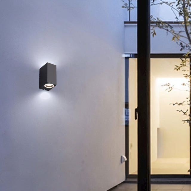 "Modern outdoor wall lamp with a dark aluminum body and glass diffuser, mounted on a contemporary building exterior, casting focused light."
