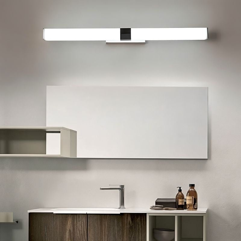"A silver wall lamp with a linear LED bar mounted above a modern bathroom vanity with a white mirror, dark wood cabinets, and toiletries."