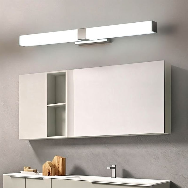 "A silver wall lamp with a linear LED bar illuminating a modern bathroom with white cabinets, a divided mirror, and decorative wooden objects."