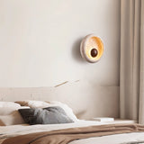 "A minimalist wall lamp with a circular travertine frame and warm wooden centerpiece serving as a sconce in a modern bedroom."