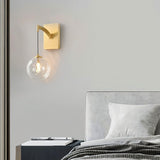"A modern brass wall lamp with a spherical, clear glass shade, mounted on a gray wall above a nightstand in a contemporary bedroom."