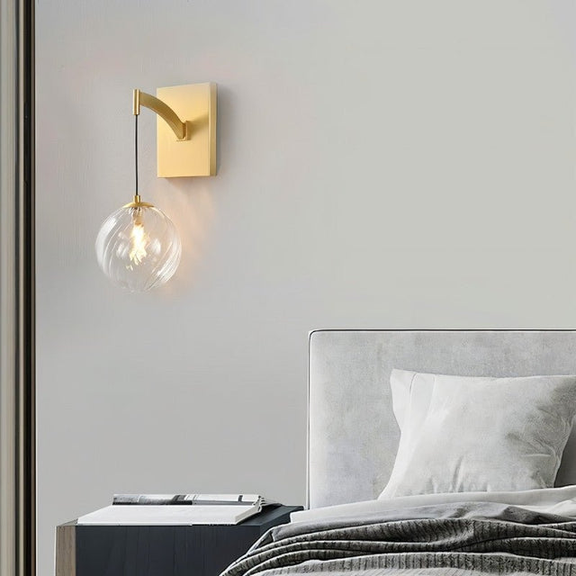 "A modern brass wall lamp with a spherical, clear glass shade, mounted on a gray wall above a nightstand in a contemporary bedroom."