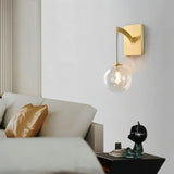 "A modern brass wall lamp with a spherical, clear glass shade with subtle ridges, illuminating a contemporary bedroom with a white wall and a comfortable sofa with a golden pillow."