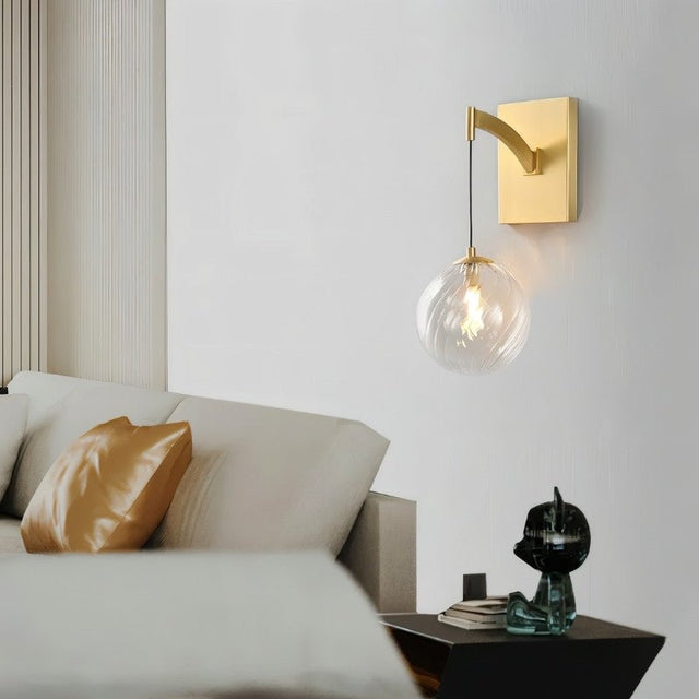 "A modern brass wall lamp with a spherical, clear glass shade with subtle ridges, illuminating a contemporary bedroom with a white wall and a comfortable sofa with a golden pillow."