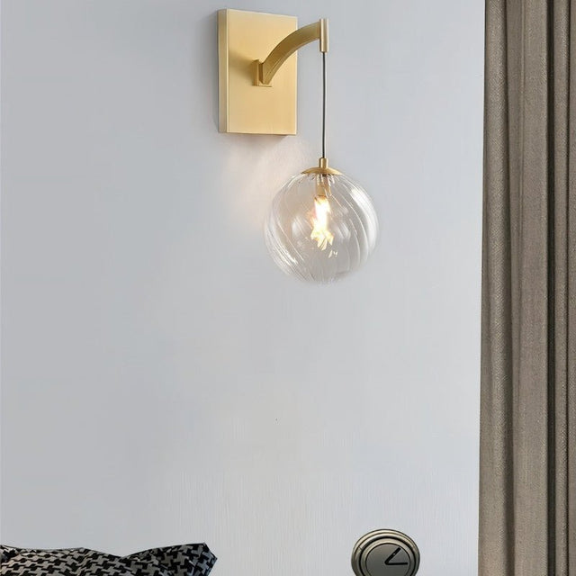 "A modern brass wall lamp with a spherical, clear glass shade with subtle ridges, providing a warm glow in a contemporary bedroom at night, mounted on a gray wall next to a window with curtains."