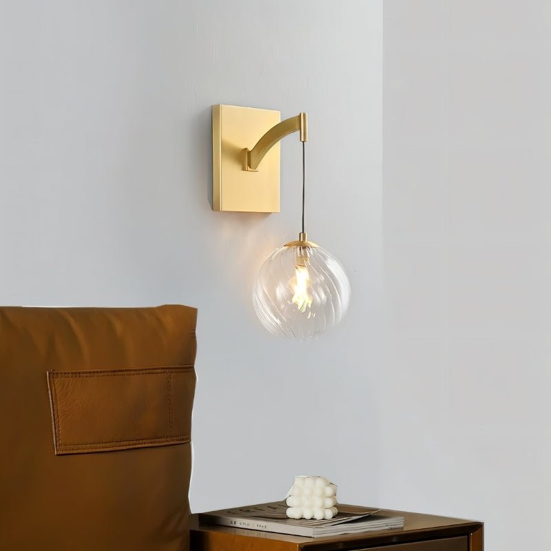 "A modern brass wall lamp with a spherical, clear glass shade with subtle ridges, providing a warm glow in a contemporary bedroom at night, mounted on a gray wall next to a leather chair and above a small table."