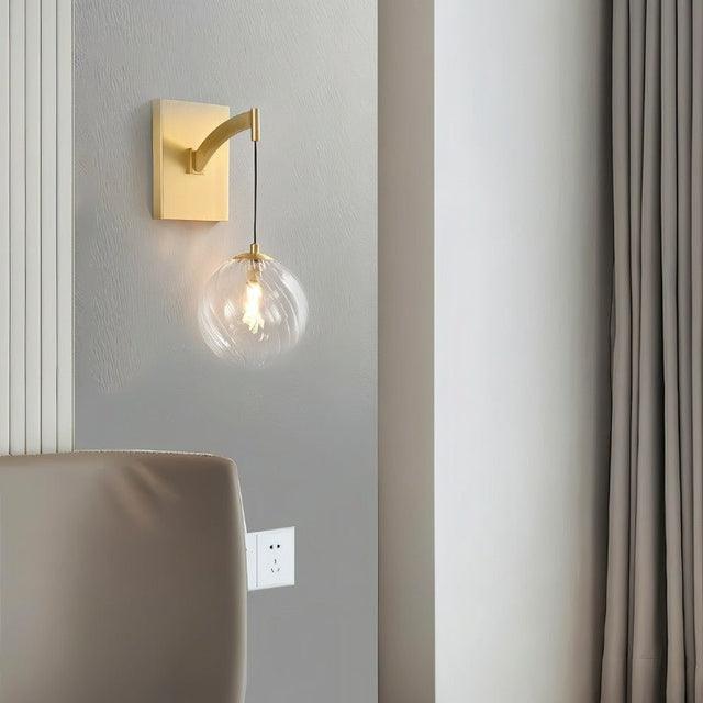 "A modern brass wall lamp with a spherical, clear glass shade with subtle ridges, mounted on a gray wall in a contemporary bedroom, illuminating the space with a warm glow."