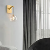 "A modern brass wall lamp with a spherical, clear glass shade with subtle ridges, mounted on a gray wall next to a window with gray and white curtains."