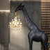 Black giraffe floor lamp holding ornate gold chandelier in modern interior with concrete walls and wooden shelving.