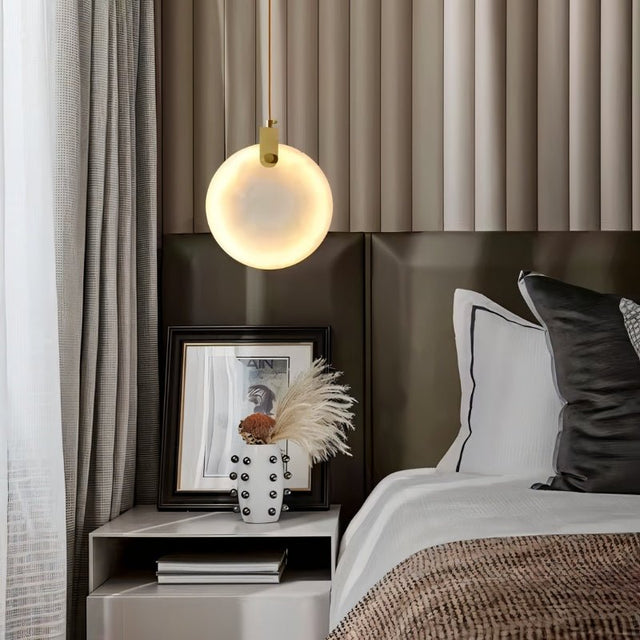 "A modern alabaster pendant light hangs above a nightstand in a contemporary bedroom, casting a warm glow."