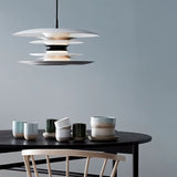 "A modern pendant light with layered discs in white and black, hanging above a dining table with cups and a chair."