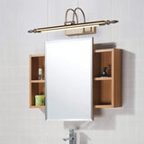 "A silver wall lamp with decorative end caps mounted above a bathroom medicine cabinet with open wooden shelves on either side, against a tiled wall."