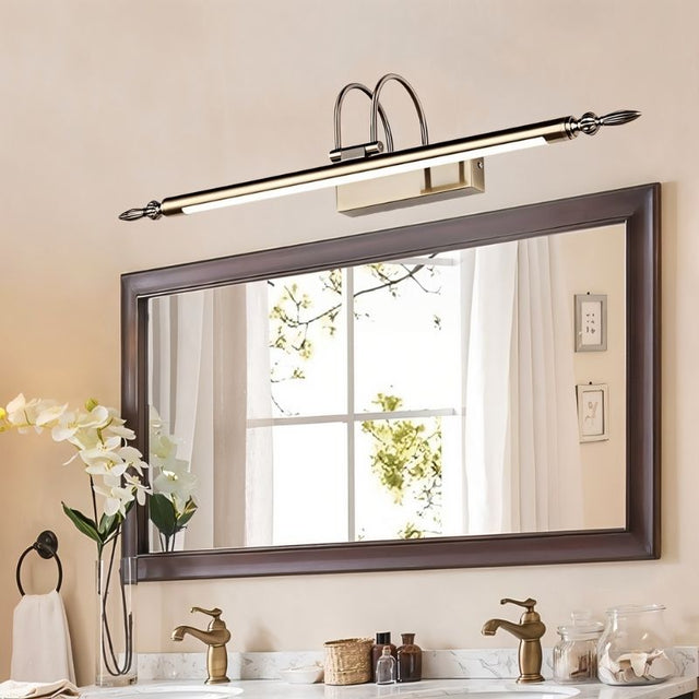"A silver wall lamp with decorative end caps mounted above a traditional framed bathroom mirror, reflecting a window with sheer curtains, with double sinks and flowers below."