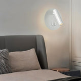 "A modern white wall-mounted reading lamp with a circular backplate and adjustable cylindrical spotlight, in a contemporary bedroom setting above a gray headboard."