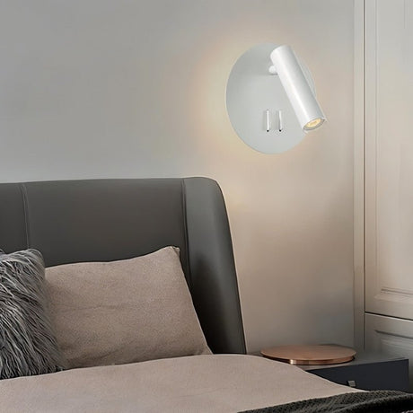 "A modern white wall-mounted reading lamp with a circular backplate and adjustable cylindrical spotlight, in a contemporary bedroom setting above a gray headboard."