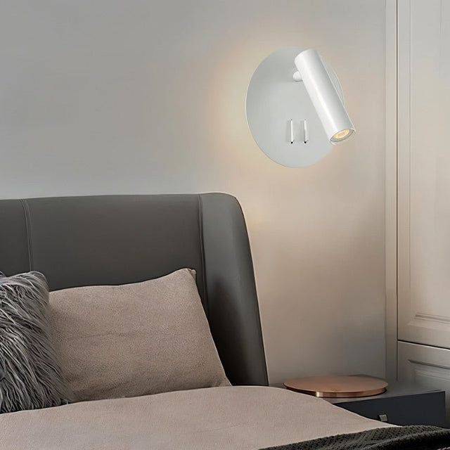 "A modern white wall-mounted reading lamp with a circular backplate and adjustable cylindrical spotlight, in a contemporary bedroom setting above a gray headboard."