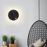 "A modern black wall-mounted reading lamp with a circular backplate and adjustable cylindrical spotlight, in a contemporary living room setting with a hanging egg chair."