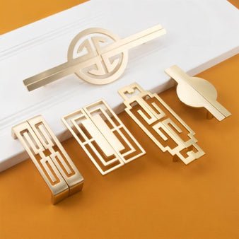 "An assortment of brushed gold, geometric pull bars with bold cutouts, made of zinc alloy, on a white and orange surface."