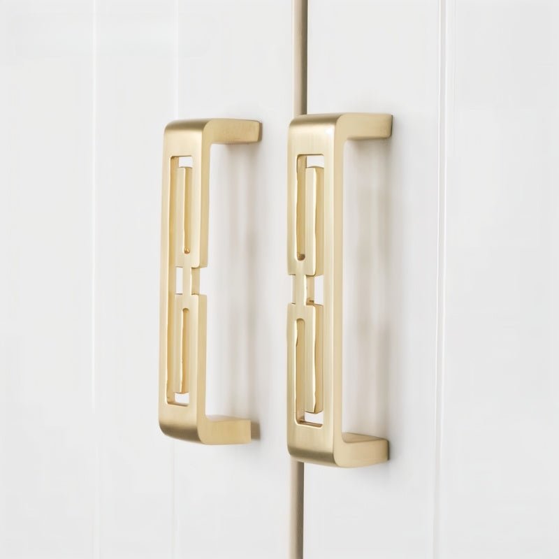"Two brushed gold, geometric pull bars with bold cutouts, made of zinc alloy, on white cabinet doors."