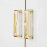 "Two brushed gold, geometric pull bars with bold cutouts, made of zinc alloy, on white cabinet doors."