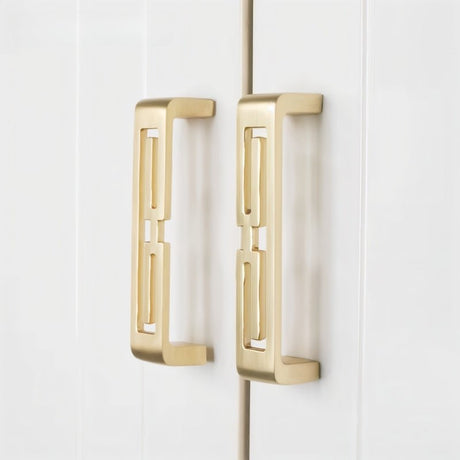 "Two brushed gold, geometric pull bars with bold cutouts, made of zinc alloy, on white cabinet doors."