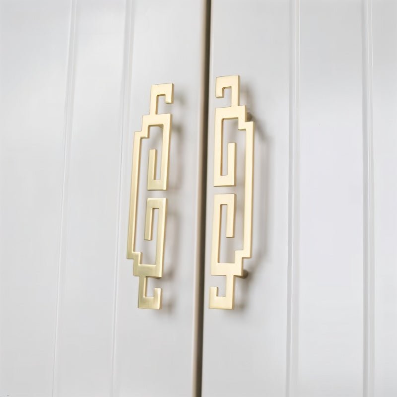 "Two brushed gold, geometric pull bars with bold cutouts made of zinc alloy on a white cabinet."