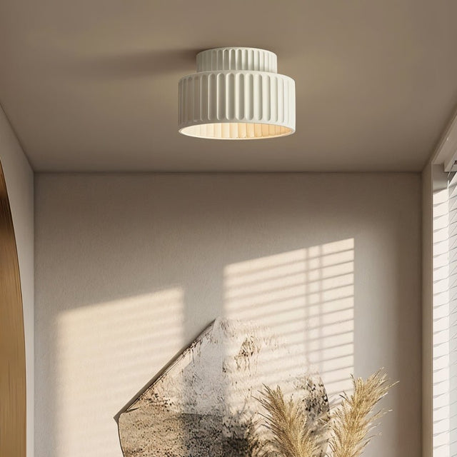 "A white ceiling light with a ribbed, cylindrical shade, shown installed on a white ceiling in a hallway with a piece of abstract art and dried plants."