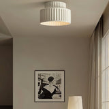 "A white ceiling light with a ribbed, cylindrical shade, shown installed on a white ceiling in a living room with a framed picture and a table lamp."